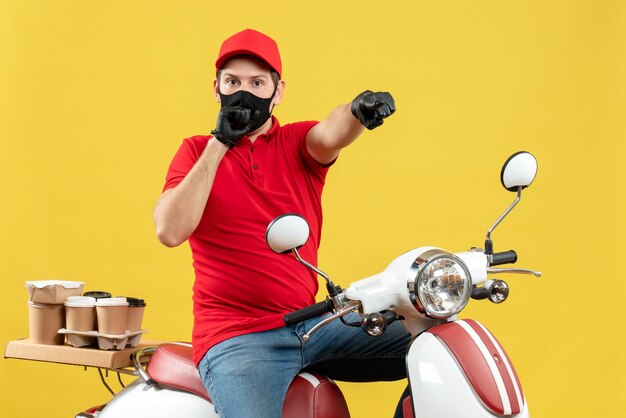 Photo gratuite vue de dessus du jeune adulte surpris portant un chemisier rouge et des gants de chapeau dans un masque médical délivrant l'ordre assis sur un scooter pointant vers l'avant sur le mur jaune