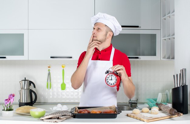 Vue de dessus du jeune chef masculin rêveur tenant une horloge dans la cuisine blanche