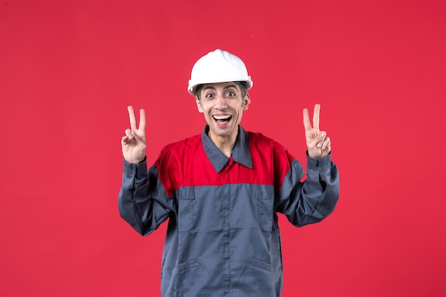Vue de dessus du jeune constructeur souriant en uniforme portant un casque de sécurité mking geste de victoire sur un mur rouge isolé