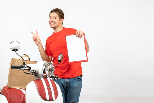 Vue de dessus du jeune livreur souriant en uniforme rouge debout près de scooter montrant le document faisant le geste de la victoire sur le mur blanc