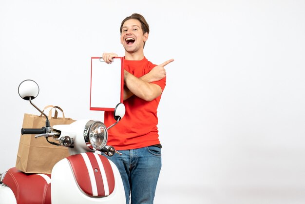 Vue De Dessus Du Jeune Livreur Souriant En Uniforme Rouge Debout Près De Scooter Montrant Le Document Et Pointant Vers L'arrière Sur Le Mur Blanc