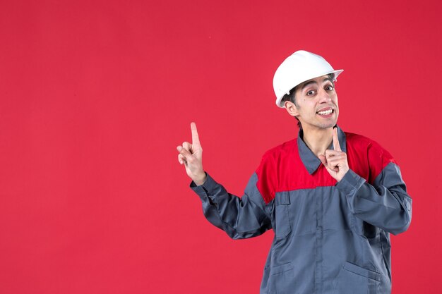 Vue de dessus du jeune travailleur curieux en uniforme dans un casque pointant vers le haut sur un mur rouge isolé
