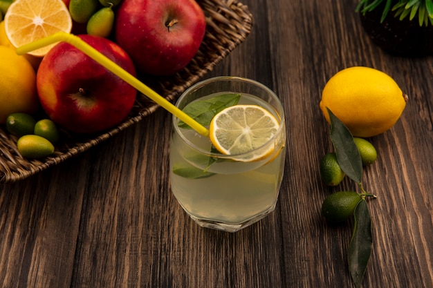 Vue de dessus du jus de citron frais avec des pommes, des citrons et des kinkans sur un plateau en osier sur un mur en bois