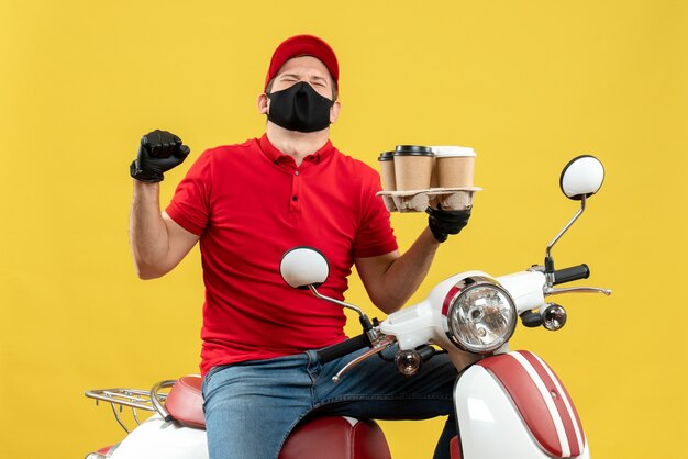 Vue de dessus du livreur émotionnel fier heureux portant des gants uniformes et chapeau en masque médical assis sur un scooter montrant les commandes