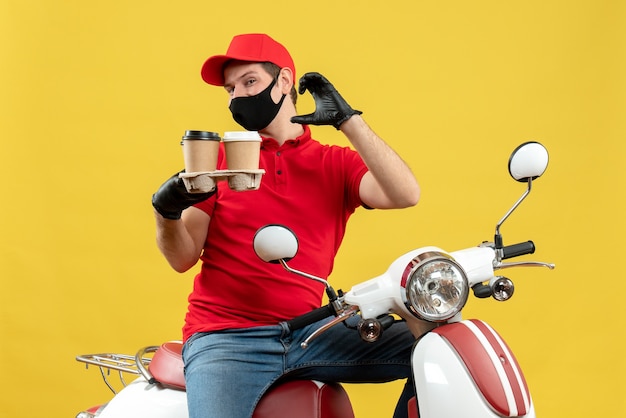 Vue de dessus du livreur fier portant des gants uniformes et chapeau en masque médical assis sur un scooter montrant des commandes faisant quelque chose d'exact