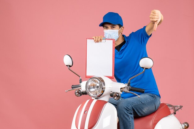 Vue de dessus du livreur masculin en masque portant un chapeau assis sur un scooter montrant un document faisant un mauvais geste sur fond de pêche pastel