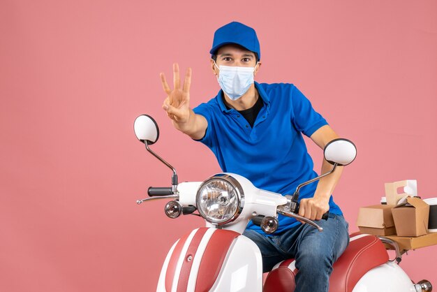 Vue de dessus du livreur en masque médical portant un chapeau assis sur un scooter et montrant trois sur fond de pêche pastel