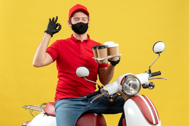 Vue de dessus du livreur de rêve portant des gants uniformes et chapeau en masque médical assis sur un scooter montrant les commandes faisant le geste de lunettes