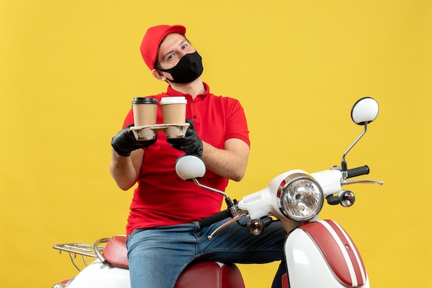 Vue de dessus du livreur souriant portant des gants uniformes et chapeau en masque médical assis sur un scooter donnant des commandes