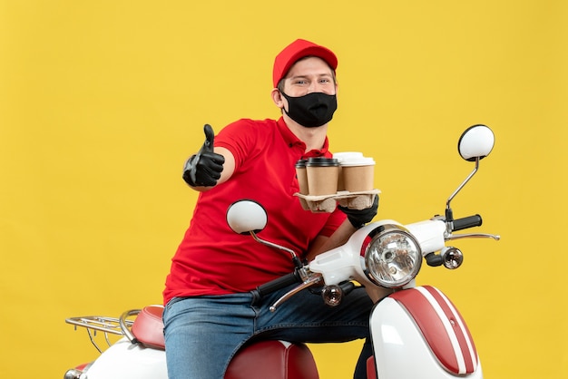 Vue de dessus du livreur souriant portant des gants uniformes et chapeau en masque médical assis sur un scooter montrant des commandes faisant un geste ok