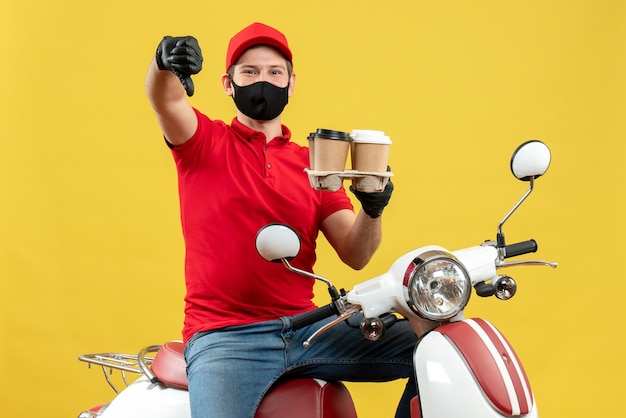 Vue de dessus du livreur souriant portant des gants uniformes et chapeau en masque médical assis sur un scooter montrant l'ordre avec le pouce vers le bas