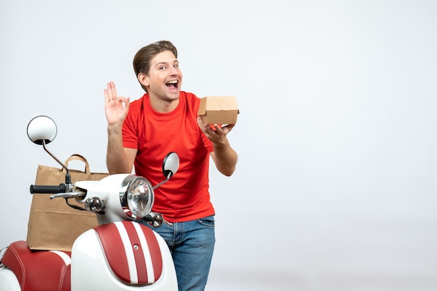 Vue de dessus du livreur souriant en uniforme rouge debout près de la commande de maintien du scooter et faisant un geste perpect sur le mur blanc