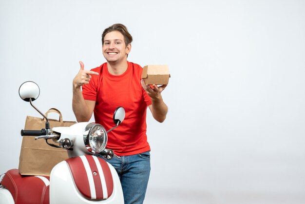 Vue de dessus du livreur souriant en uniforme rouge debout près de l'ordre de pointage du scooter sur le mur blanc