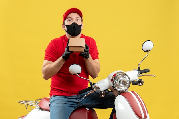 Vue de dessus du livreur surpris portant un chemisier rouge et des gants de chapeau en masque médical assis sur un scooter montrant l'ordre