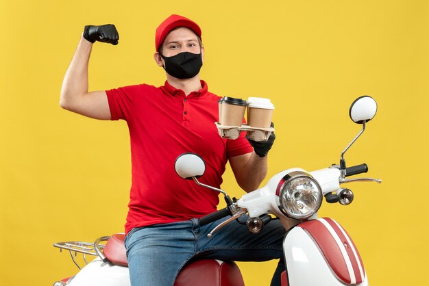 Vue de dessus du livreur surpris portant des gants uniformes et chapeau en masque médical assis sur un scooter tenant des commandes montrant son musclé