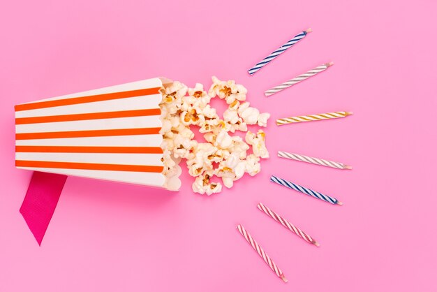 Une vue de dessus du maïs soufflé frais à l'intérieur du paquet de papier isolé avec des bougies colorées sur rose, collation de graines de maïs film