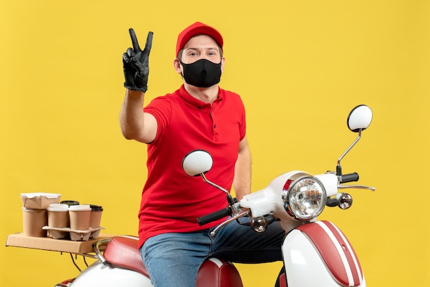 Vue de dessus du mec de messagerie souriant portant chemisier rouge et gants de chapeau en masque médical délivrant l'ordre assis sur scooter faisant le geste de la victoire