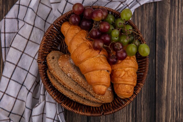 Vue de dessus du pain comme des croissants et des tranches de pain d'épi brun épépiné avec du raisin dans le panier sur un tissu à carreaux sur fond de bois