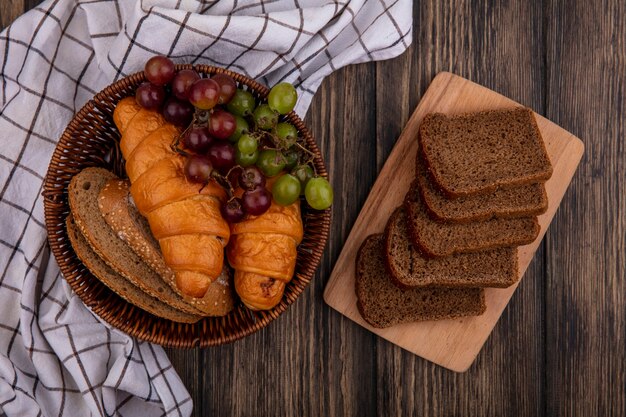 Vue de dessus du pain comme des croissants et des tranches de pain d'épis brun épépiné avec du raisin dans le panier sur un tissu à carreaux et des tranches de pain de seigle sur une planche à découper sur fond de bois