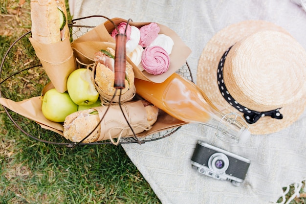 Vue de dessus du panier avec pommes, pain et bouteille de jus d'orange. Photo d'en haut de la nourriture pour le déjeuner, appareil photo et chapeau de paille allongé sur une couverture blanche sur l'herbe.