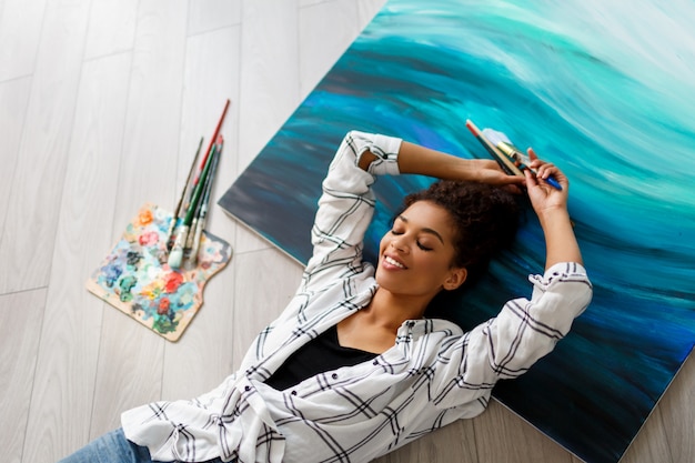 Vue de dessus du peintre heureux Africana femme américaine couchée sur toile et regardant la caméra avec des pinceaux dans les mains.