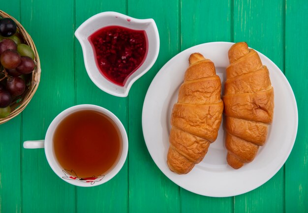 Vue de dessus du petit-déjeuner avec des croissants en assiette tasse de thé confiture de framboises dans un bol et panier de baies de prunelle de raisin sur fond vert