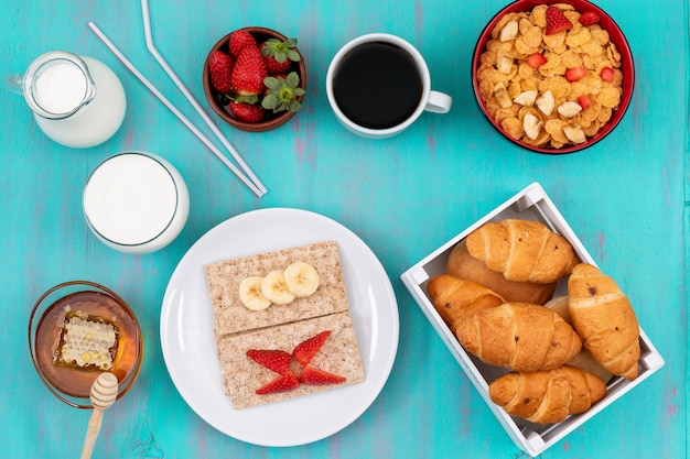Vue de dessus du petit déjeuner avec croissants, cornflakes, fruits, lait et miel sur la surface bleue horizontale