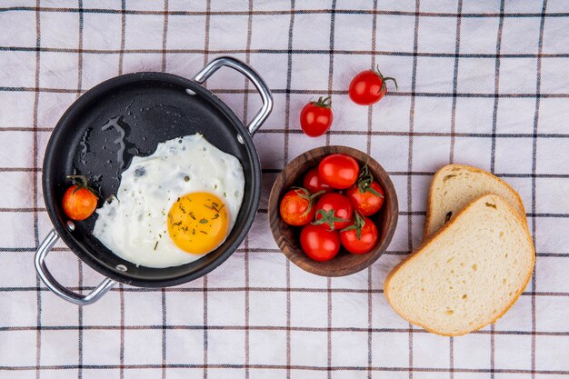 Vue de dessus du petit-déjeuner avec poêle d'oeuf au plat et bol de tomate avec des tranches de pain sur un tissu à carreaux