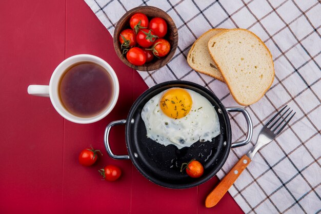 Vue de dessus du petit-déjeuner avec poêle d'oeuf au plat et tomate avec bol de tranches de pain de tomate fourchette sur tissu à carreaux et tasse de thé sur rouge