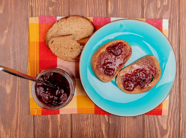 Vue de dessus du petit-déjeuner avec des tranches de pain de seigle enduites de confiture dans une assiette et des morceaux de pain de seigle confiture de fraises sur tissu à carreaux et table en bois