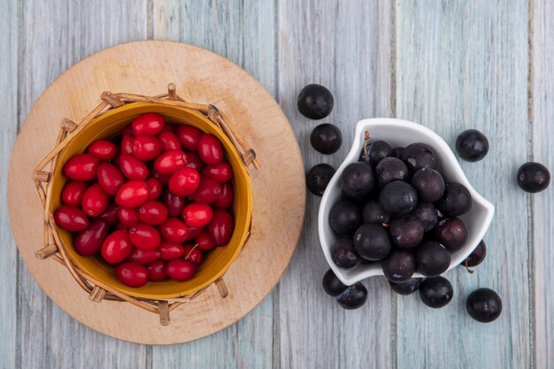 Vue de dessus du petit prunellier violet foncé sur un bol blanc avec des baies de cornouiller rouge sur un seau sur une planche de cuisine en bois sur un fond en bois gris