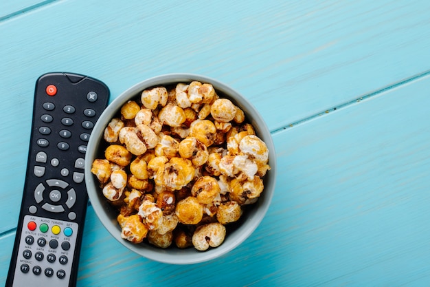 Vue de dessus du pop-corn au caramel doux dans un bol et une télécommande de télévision sur fond bleu