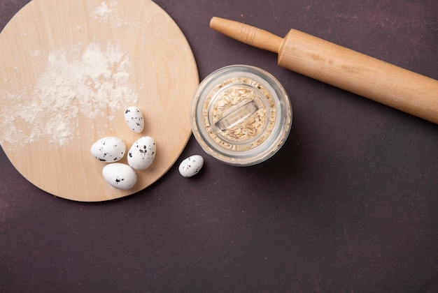 Vue de dessus du pot plein de flocons d'avoine et rouleau à pâtisserie avec des œufs et de la farine sur une planche à découper sur fond marron avec copie espace