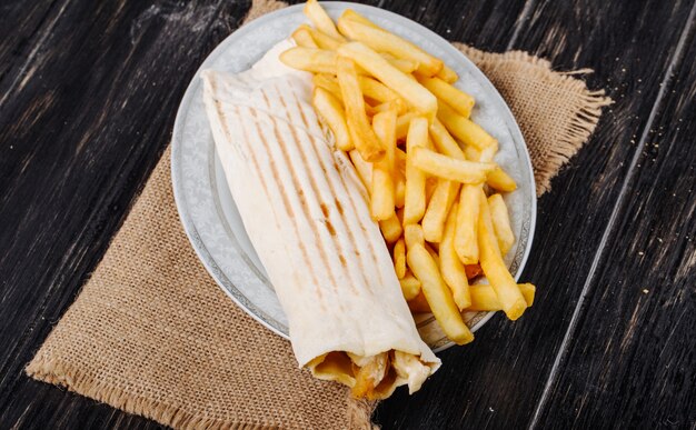 Vue de dessus du poulet doner enveloppé dans du lavash servi avec des frites sur une assiette sur un sac sur fond rustique