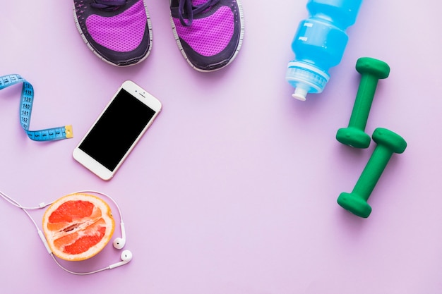 Vue de dessus du ruban à mesurer; haltère; chaussures; fruits orange coupés en deux; bouteille d&#39;eau; téléphone portable et écouteurs sur fond rose