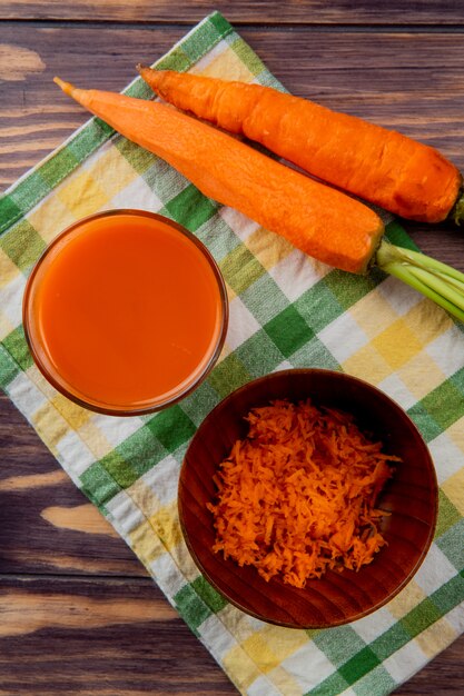 Vue de dessus du verre de jus de carotte avec bol de carotte râpée et carottes entières sur tissu sur fond de bois