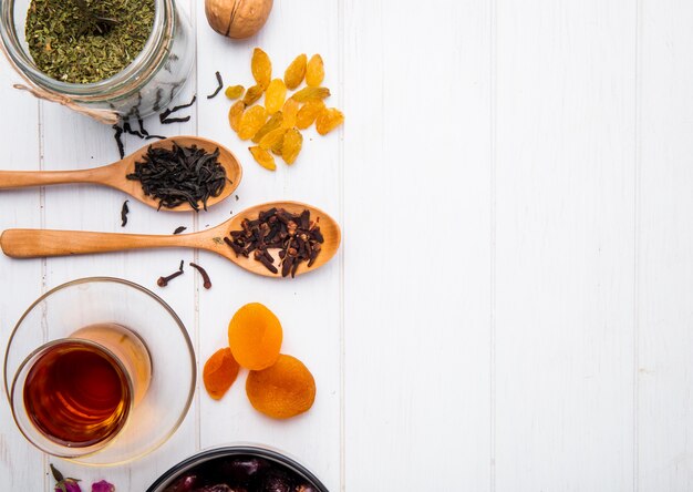 Vue de dessus du verre de thé armudu avec des abricots secs secs et des raisins secs et des cuillères en bois avec des feuilles de thé noir sec et des épices de clou de girofle sur du bois blanc avec copie espace