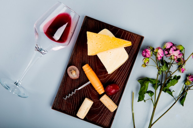 Vue de dessus du verre de vin rouge avec des fleurs et du cheddar de raisin et des bouchons de fromage parmesan et tire-bouchon sur une planche à découper sur le tableau blanc