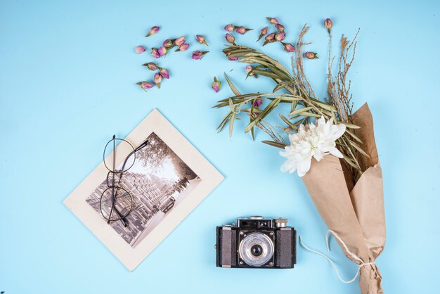 Vue de dessus du vieil appareil photo avec une fleur de chrysanthème de couleur blanche dans du papier kraft et des boutons de rose secs éparpillés sur bleu