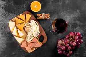 Photo gratuite vue de dessus du vin rouge avec raisin, orange et fromage sur une planche à découper en bois sur une surface sombre horizontale