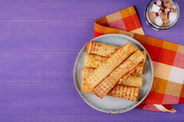 Photo gratuite vue de dessus du wafer roll rempli de lait concentré sur une plaque et des morceaux de sucre dans un bocal en verre sur fond violet en bois