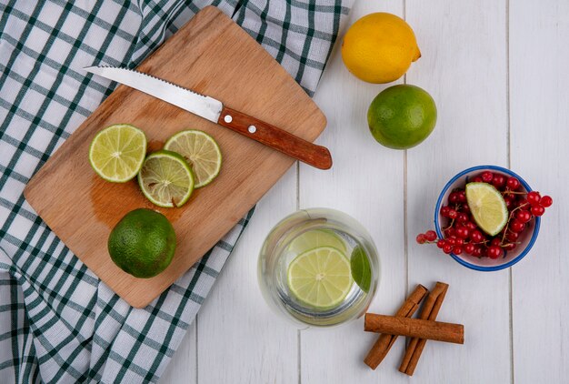 Vue de dessus de l'eau dans un verre avec de la chaux et du citron sur une planche avec un couteau et de la cannelle sur un tableau blanc