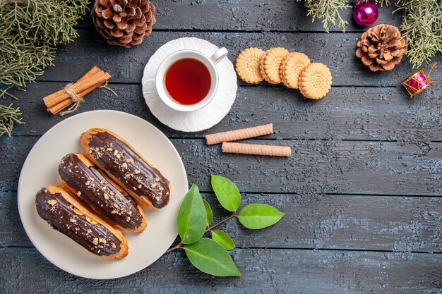 Vue de dessus éclairs au chocolat sur cônes de plaque ovale blanche jouets de Noël sapin feuilles de cannelle et différents biscuits sur un sol en bois foncé avec des espaces de copie