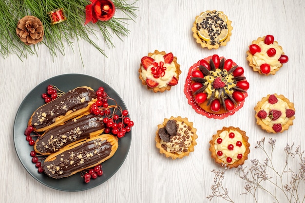 Vue de dessus éclairs au chocolat et groseilles sur la plaque grise gâteau aux baies arrondi avec des tartes et des feuilles de pin avec des jouets de Noël sur le sol en bois blanc