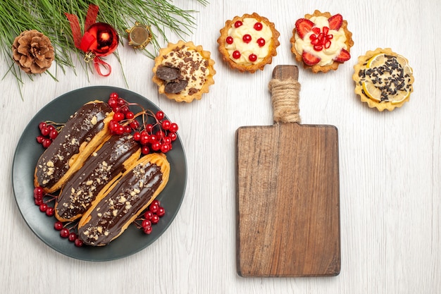 Vue de dessus éclairs au chocolat et groseilles sur la plaque grise tartes une planche à découper et des feuilles de pin avec des jouets de Noël sur le sol en bois blanc