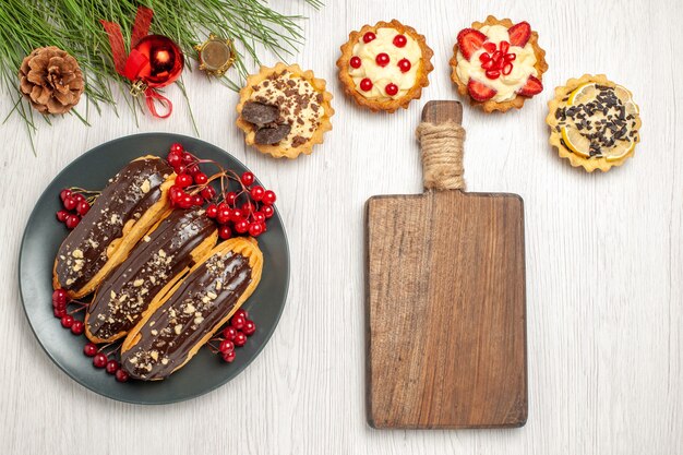 Vue de dessus éclairs au chocolat et groseilles sur la plaque grise tartes une planche à découper et des feuilles de pin avec des jouets de Noël sur le sol en bois blanc