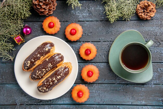 Vue de dessus éclairs au chocolat sur plaque ovale blanche branches de sapin jouets de Noël cupcakes et une tasse de thé sur une table en bois foncé