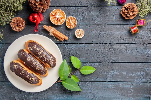 Vue de dessus éclairs au chocolat sur une plaque ovale blanche cônes jouets de Noël sapin feuilles cannelle séchée orange sur un sol en bois foncé avec copie espace