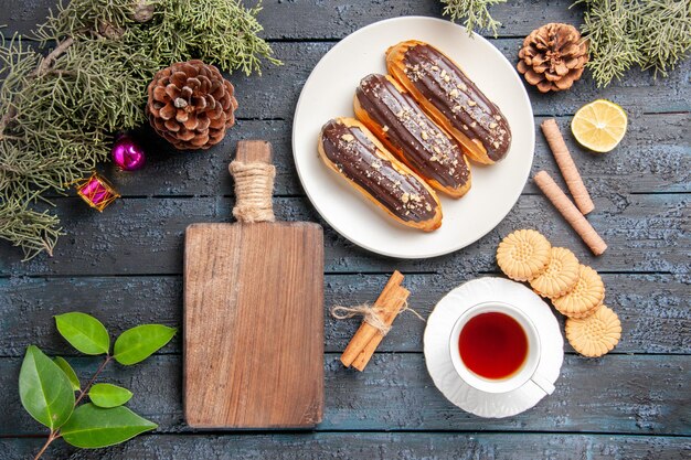 Vue de dessus éclairs au chocolat sur plaque ovale blanche cônes de sapin feuilles de cannelle tranche de citron différents biscuits une tasse de thé et une planche à découper sur un sol en bois foncé