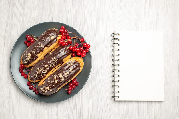 Vue de dessus des éclairs au chocolat et des raisins de Corinthe sur la plaque grise et un cahier sur la table en bois blanc